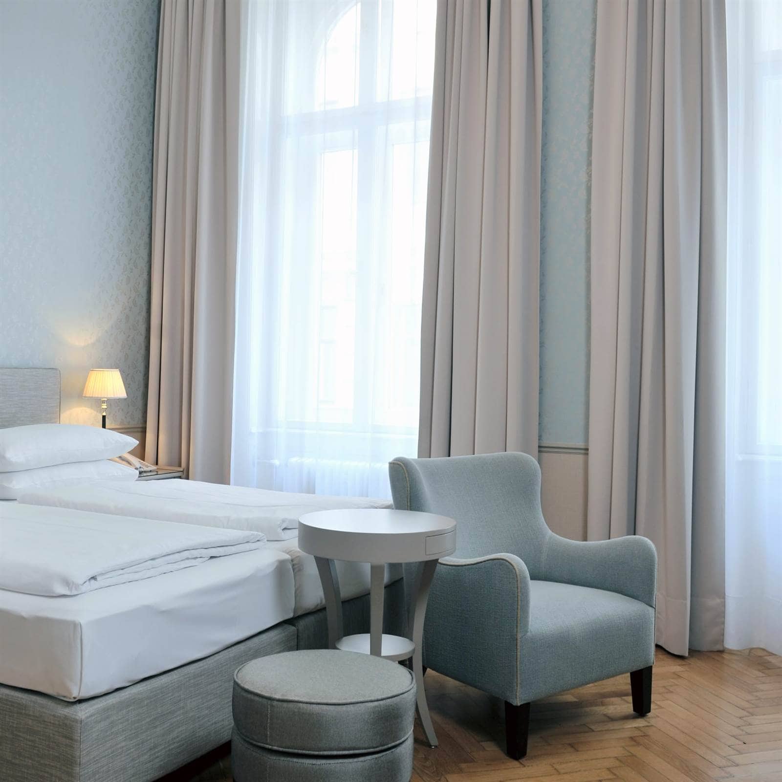 Doppelzimmer Medium im Hotel Kärntnerhof mit Bett, hellblauem Fauteuil und rundem Tisch vor dem hohen Fenster