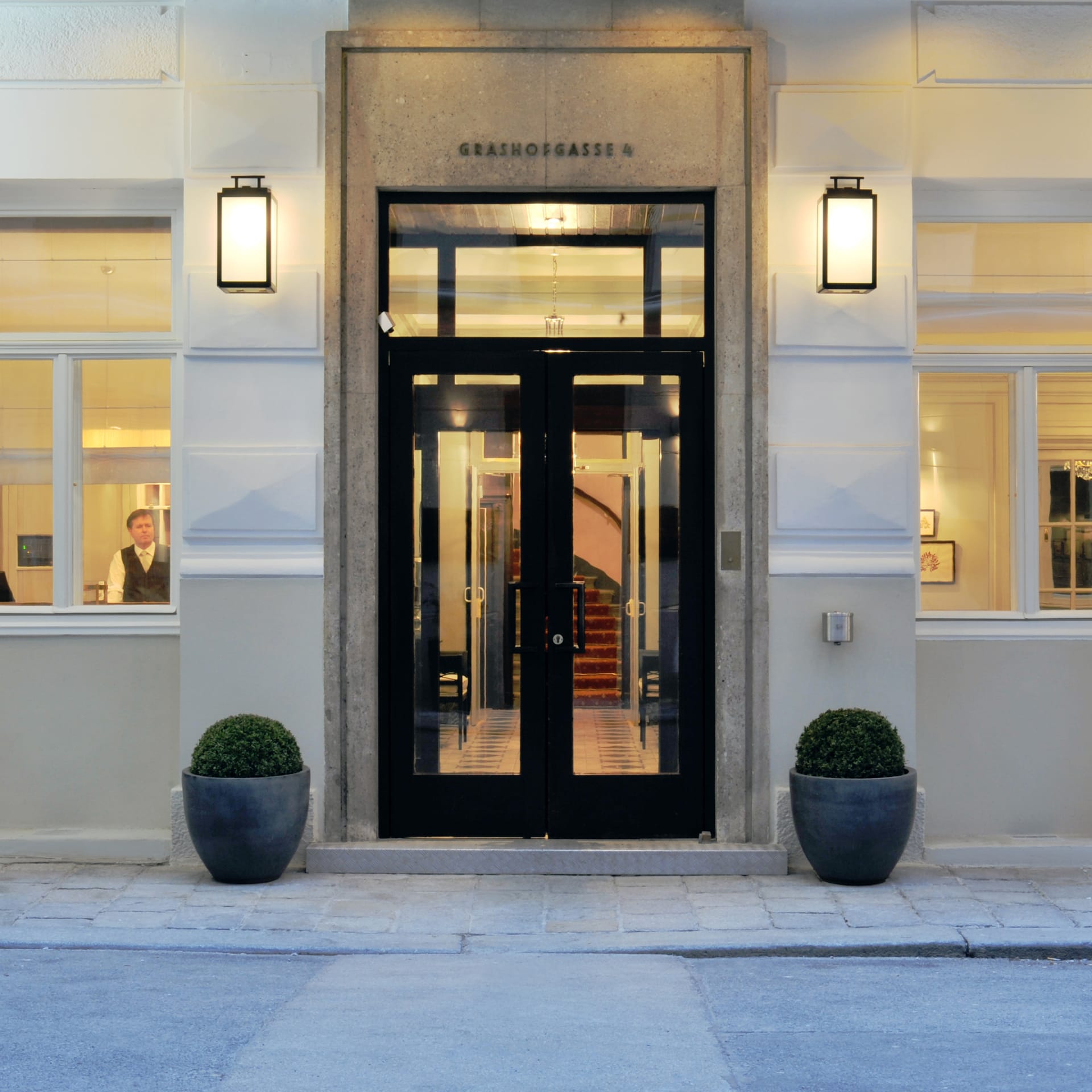 Beleuchteter Eingang des Hotel Kärntnerhof in der Grashofgasse 4 in Wien am Abend, schwarze Glastür zwischen zwei Laternen, davor zwei Buchsbaumkugeln