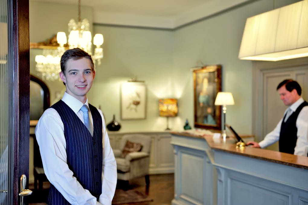 Zwei Mitarbeiter des Hotel Kärntnerhof an der Rezeption, im Hintergrund Luster, Fauteuil und gerahmte Bilder im Salon