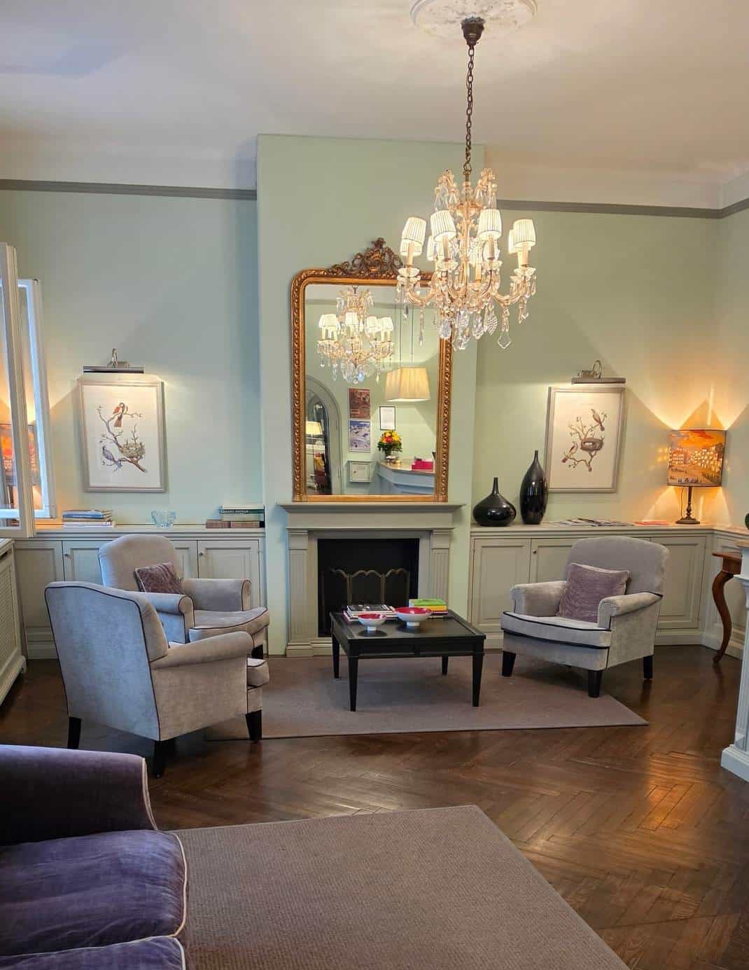Salon im Hotel Kärntnerhof mit zwei hellblauen Fauteuils, Kamin, goldgerahmtem Spiegel und Kronleuchter über Fischgrätparkett